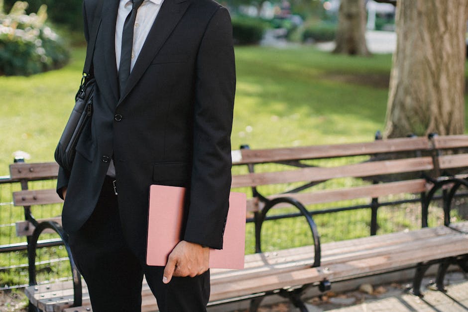 A well-dressed professional holding a folder stands near a bench in a park setting.